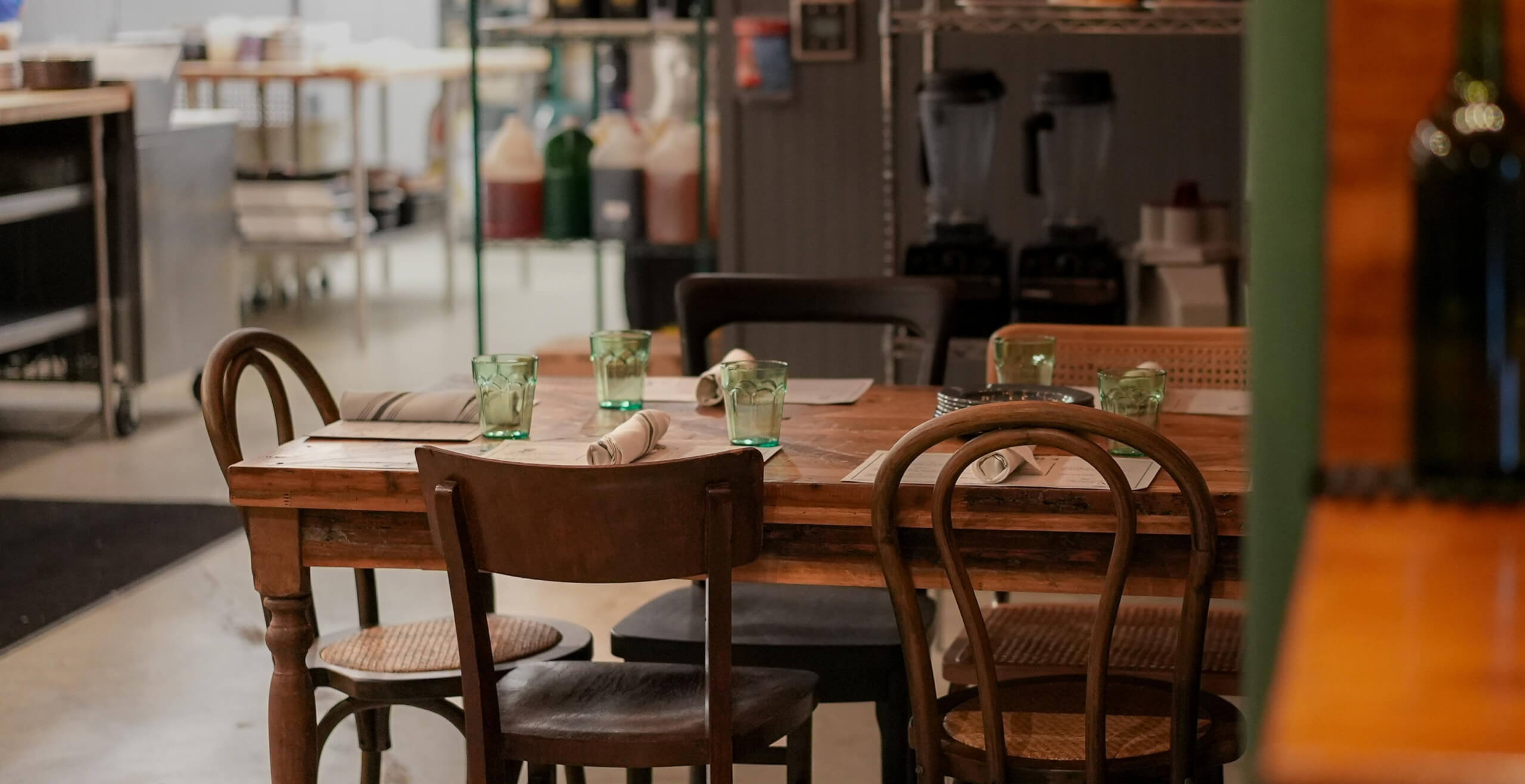 An image of a wooden table in a restaurant’s kitchen. The table has 5 place settings on it each with a green glass.