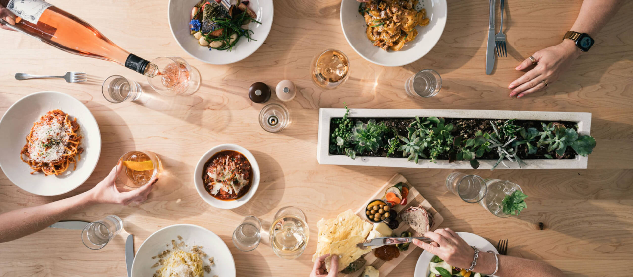 An overhead view of four people’s hands reaching for various items on a wooden table. In the lower left, someone smears a spread from a charcuterie board on a crispy cracker. In the upper right, someone is eating from a plate of pasta with a fork. In the upper left, someone is cutting into a piece of salmon while their wine glass is being refilled by a someone pouring rose. In the lower-left someone is holding an Old Fashioned in front of a plate of pasta they were just served.