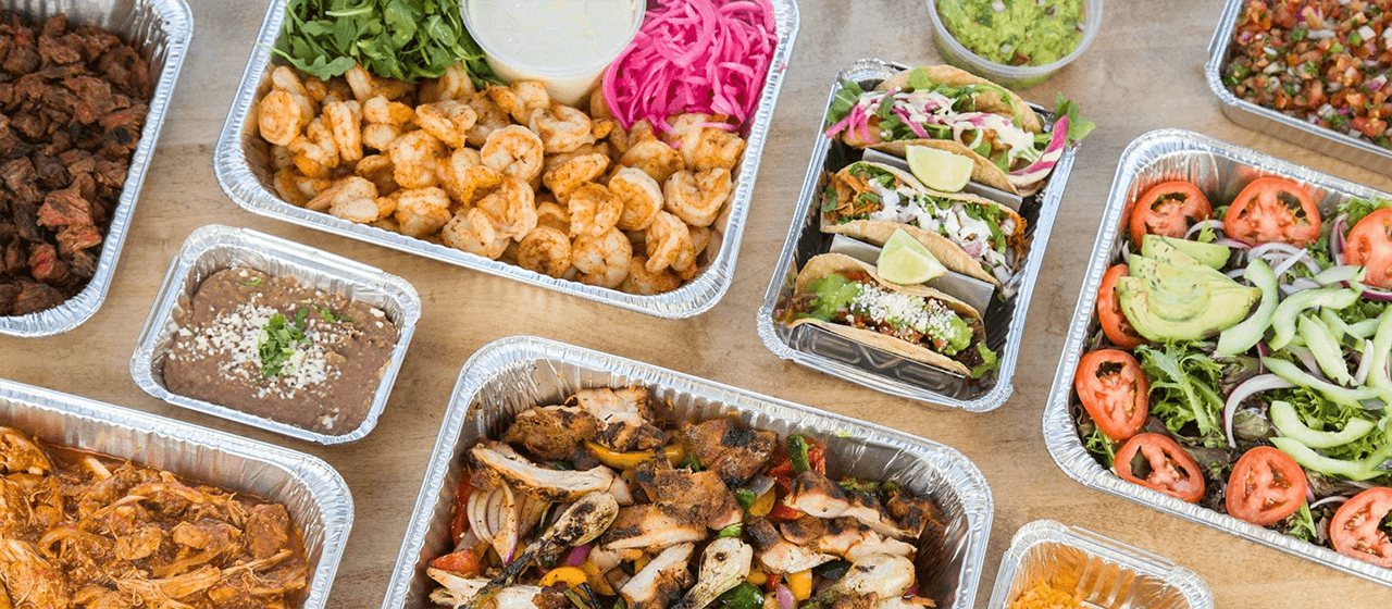 An image of takeout containers sitting on the lawn. One container has a salad, one has lamb chops, and there is a pile of pita bread with a container of tzatziki to the right.