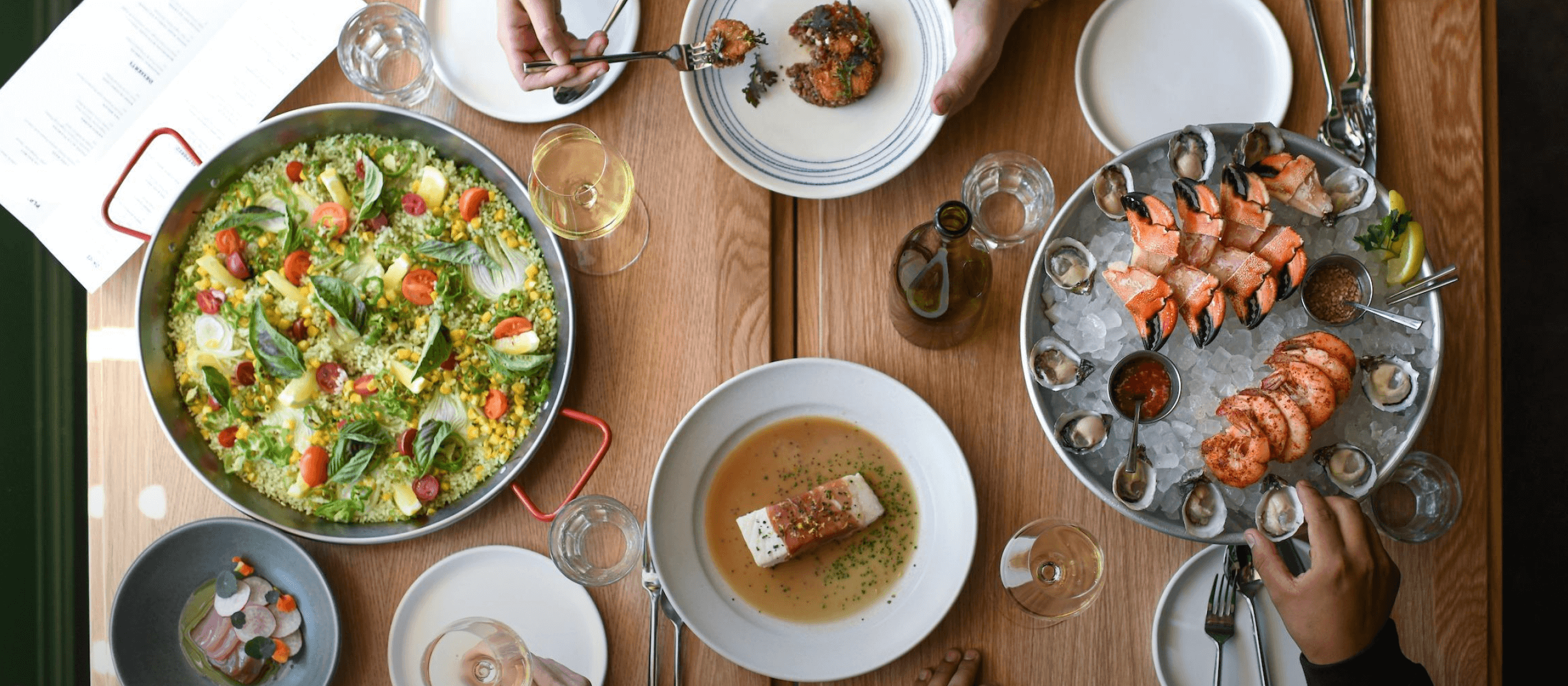 An overhead view of a table of people sharing dishes of crab claws, shrimp, oysters, fish, and paella. Glasses of wine are spread throughout the table.