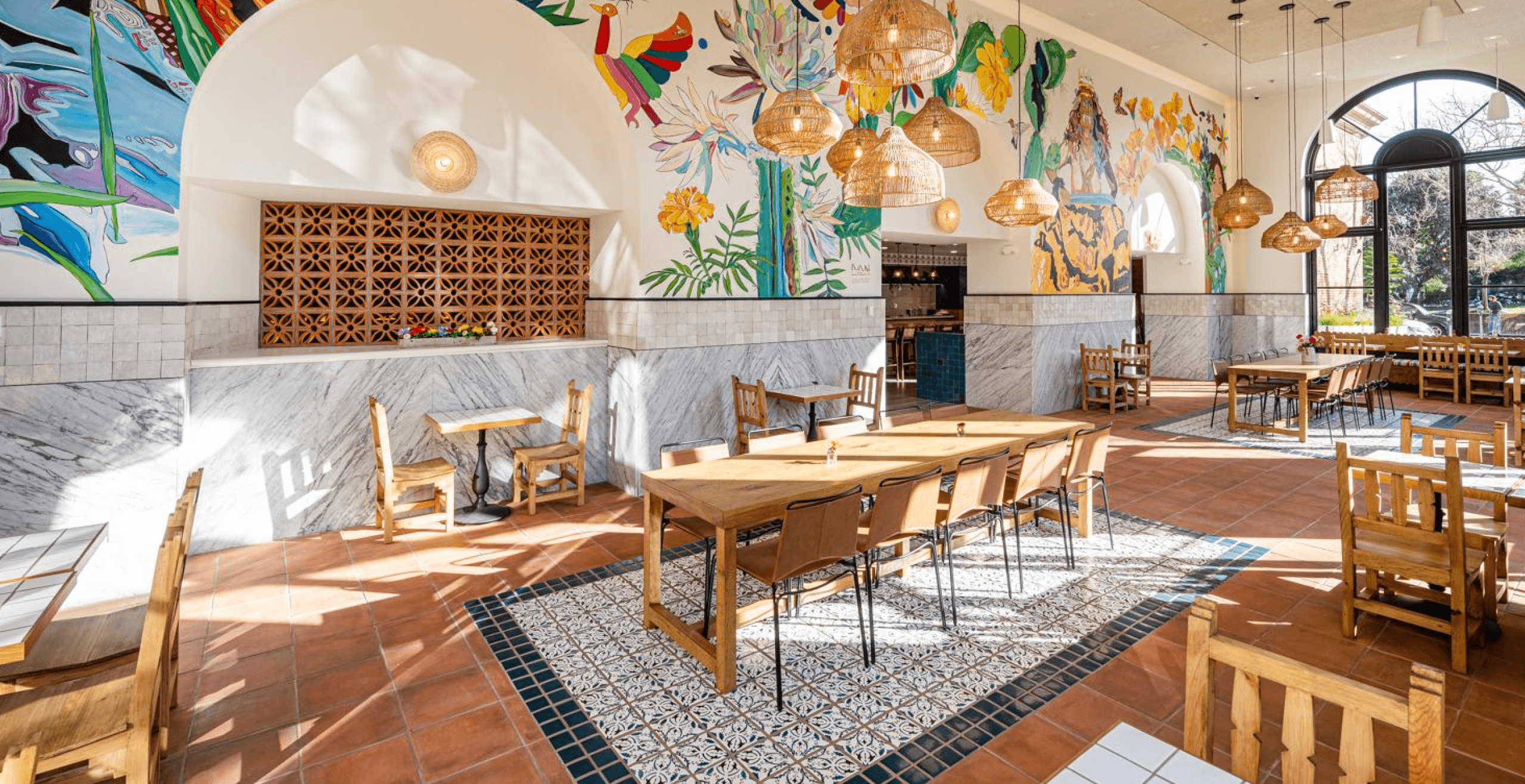 An image of a sunny dining room with wood and tiled tables and a large colorful mural on the wall.