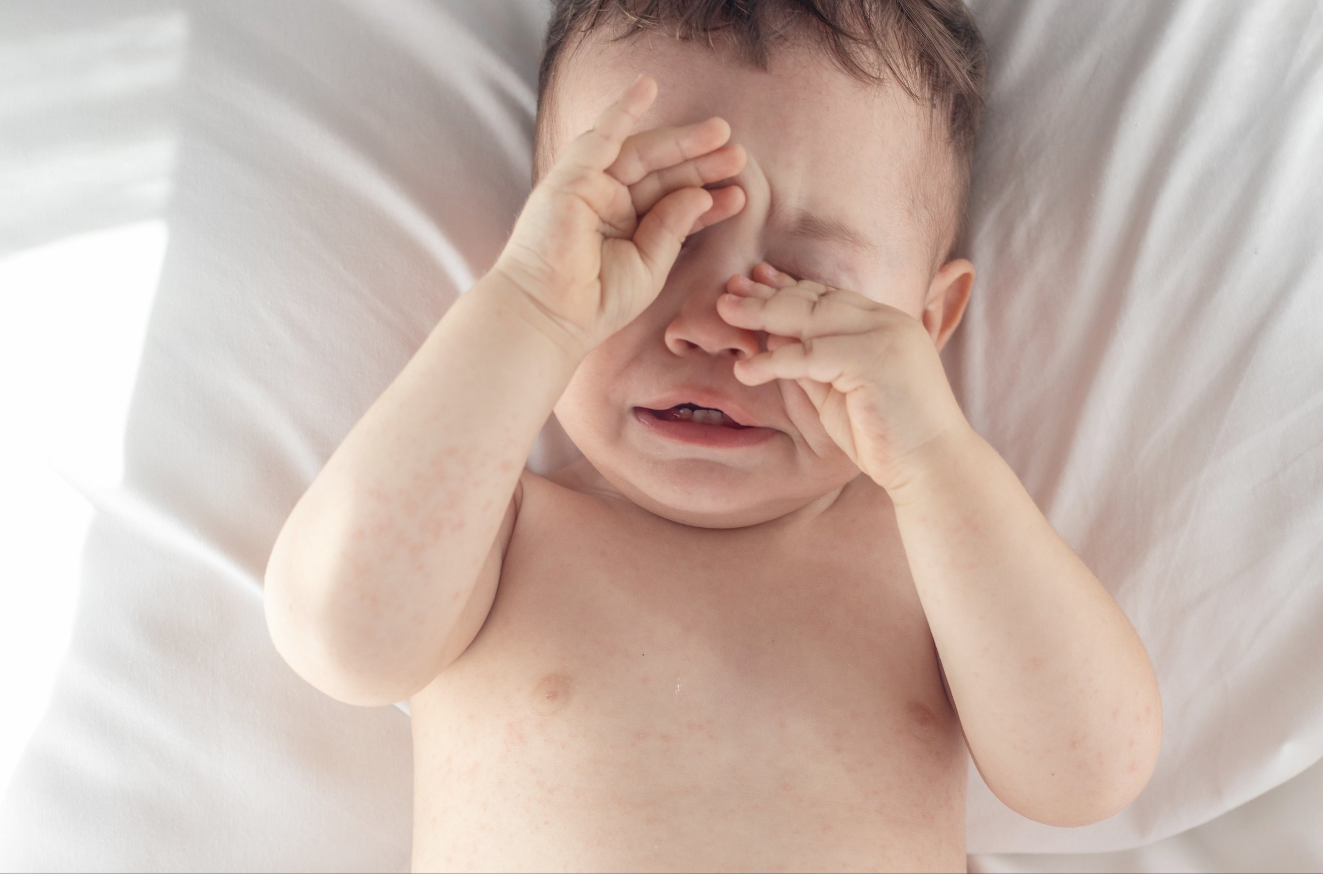 baby boy lies lying in crib on white blanket crying holding hand on eyes sleepy nervous infant