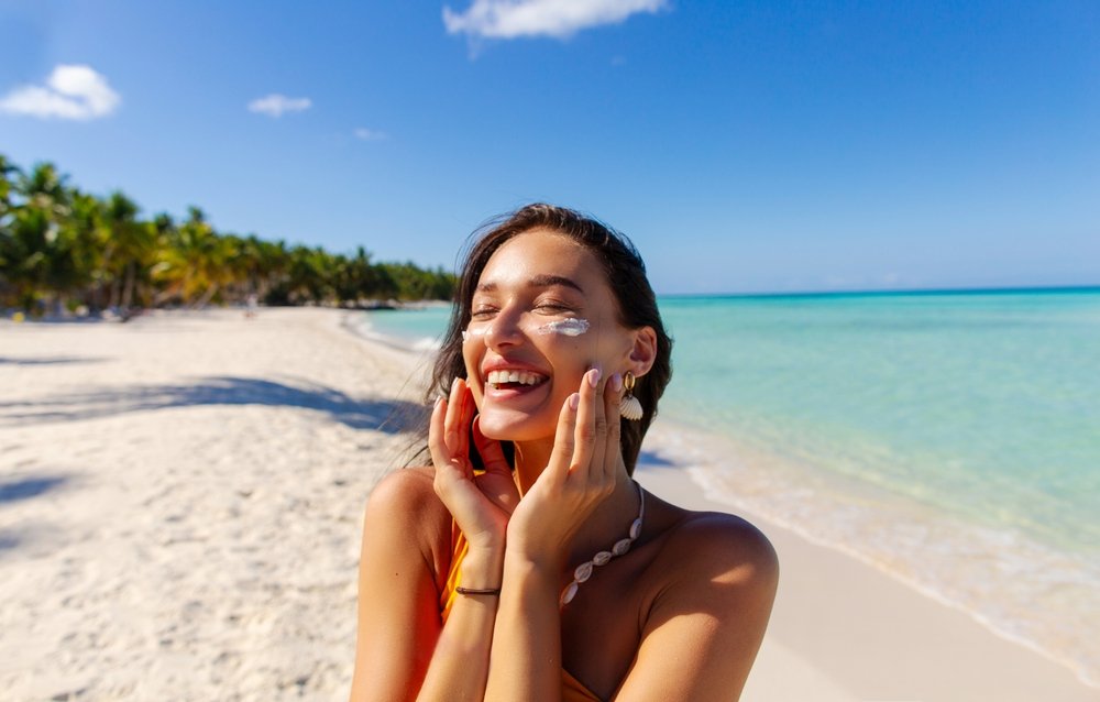 Safe sunbathing. Happy woman applying suntan lotion on face for UV-protection, standing on sandy island beach, copy space