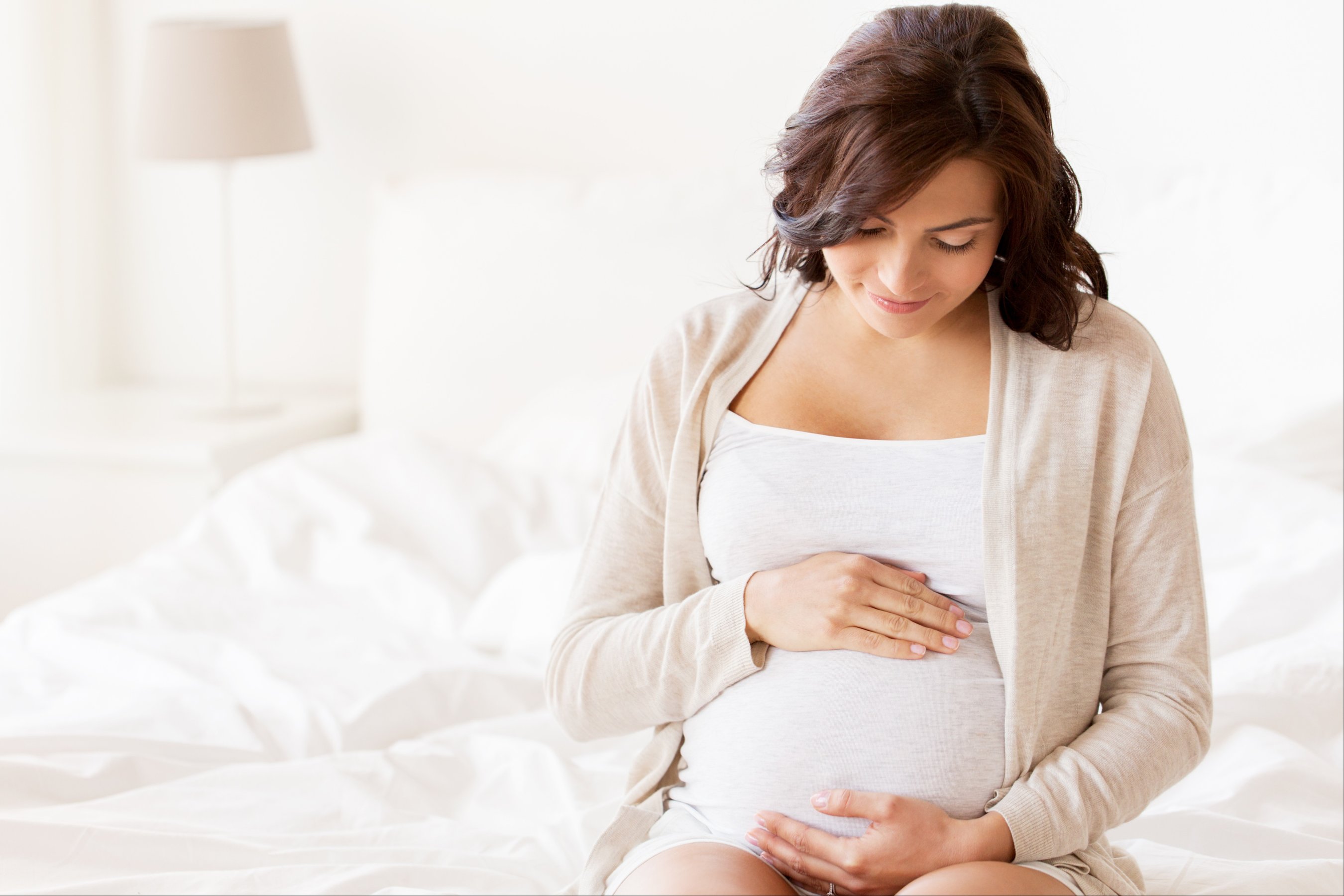 happy pregnant woman sitting on bed and touching her belly at home