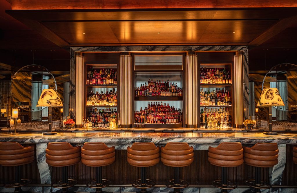 Straight view of Bar Tre Dita bar top seating. Large, dramatic marble bar filled with various Italian liquors and spirits. Pairs of brown leather barstool seats and two large patterned lamps on the two ends of the bar