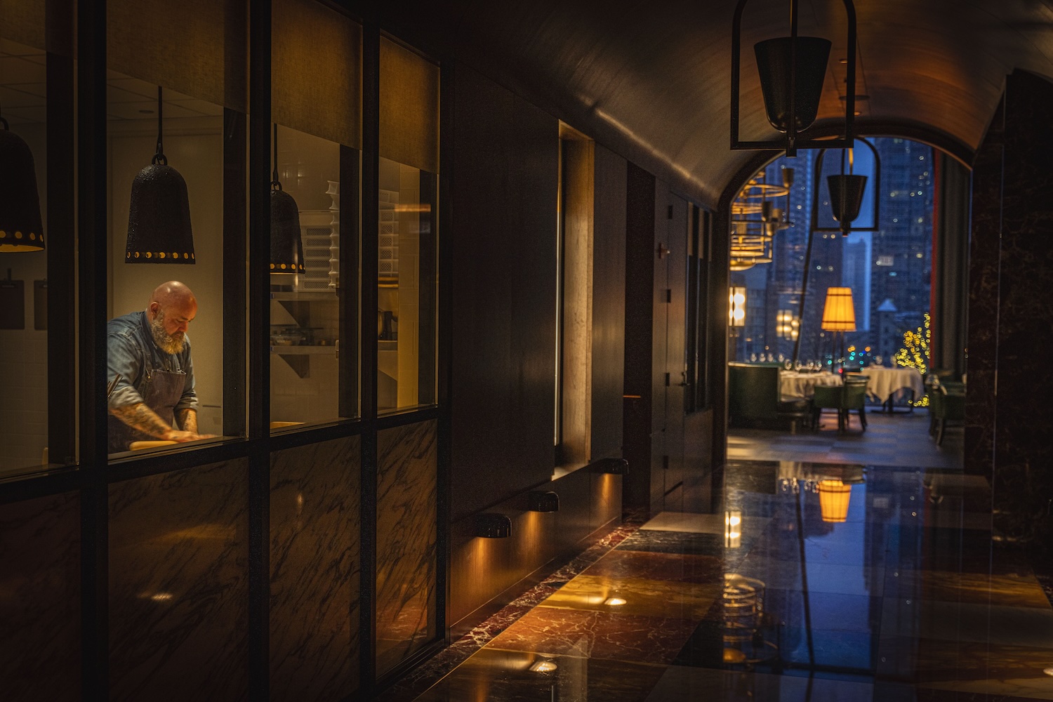 Chef Evan Funke viewed from the hallway in the pasta lab rolling pasta, with a view of the main dining room lit up at dusk in the background