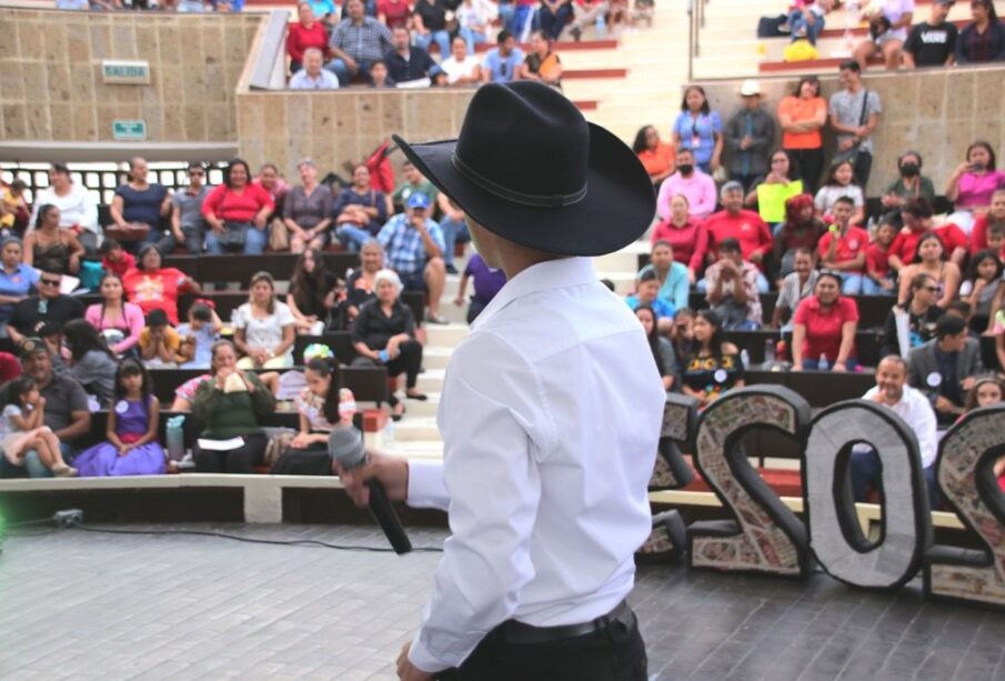 Una persona con sombrero sosteniendo un micrófono frente a personas en una explanada