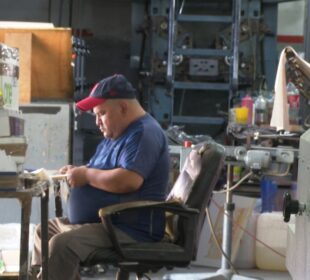 La imagen muestra a un hombre poniéndole hilo a unas etiquetas en una imprenta. El hombre está usando una máquina para colocar el hilo en las etiquetas, y las etiquetas están en un papel blanco.