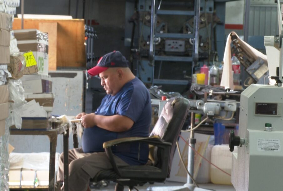 La imagen muestra a un hombre poniéndole hilo a unas etiquetas en una imprenta. El hombre está usando una máquina para colocar el hilo en las etiquetas, y las etiquetas están en un papel blanco.