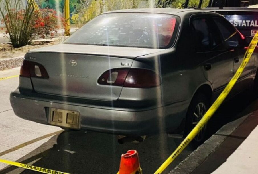 La imagen muestra un vehículo tipo Tsuru sin placas, frente a una patrulla de la policía municipal. El vehículo está rodeado de una cinta de prohibido el paso y se ven conos rojos que sostienen la cinta.