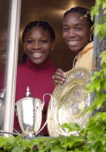 Venus Williams, right, displays the Women's Singles trophy and her sister Serena displays the Women's Doubles trophy from a window in the women's locker room in the Centre Court complex at Wimbeldon on Monday
