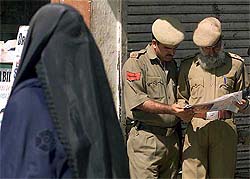 A veiled Kashmiri woman walks past policemen reading a newspaper