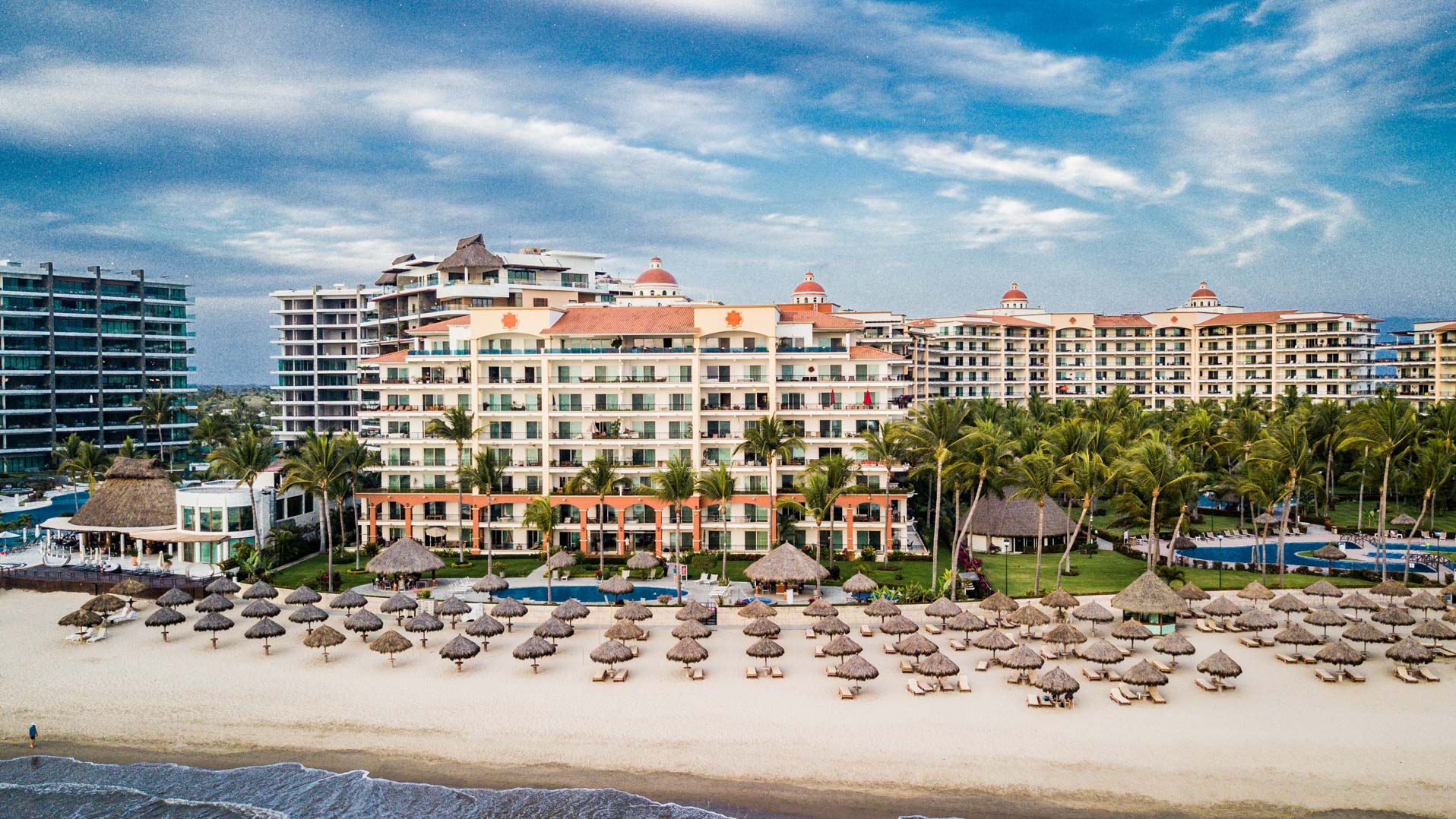 An awe-inspiring aerial photograph of Playa Royale Residences on a sun-drenched day. This captivating image offers a bird's-eye view of the resort, showcasing its proximity to the glistening white sand beaches. Beach chairs and colorful umbrellas dot the shoreline, providing shade for guests seeking refuge from the sun's rays