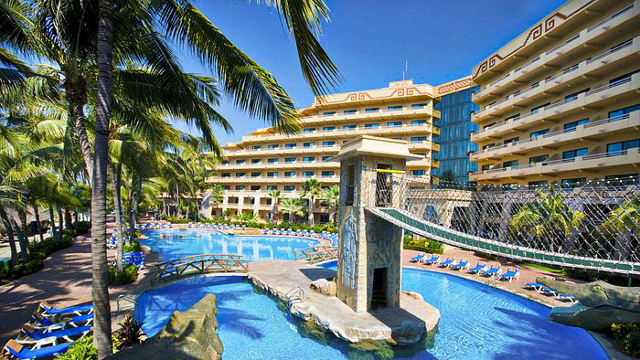 A picturesque view of Paradise Village, a luxury resort located just north of Puerto Vallarta. The image captures a beautiful, sun-filled day showcasing the resort's stunning exterior. In the foreground, there's a pristine pool surrounded by an array of comfortable sunbathing chairs, inviting guests to relax and soak up the sun