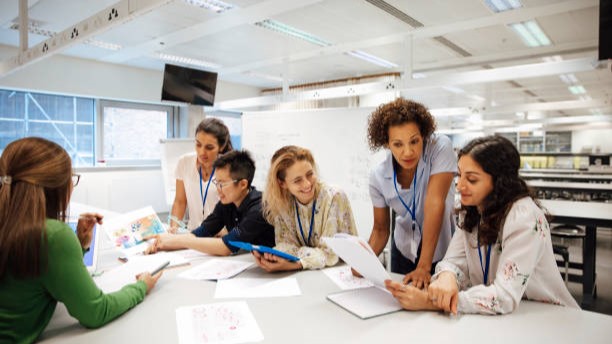 Teacher with a group of university students, in a laboratory classroom. The instructor is considering one of the students work, the mood is light hearted and positive. Other classmates are discussing things with each other. This is a realistic teaching scenario, with candid expressions. This is a multi-ethnic group of women. In the background there is a white board with mathematical formula written on it. All ladies are wearing id tags.