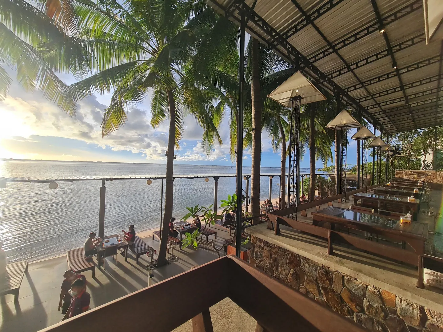 A group of people are dining at a beachfront restaurant during sunset, with a tree and lights providing an intimate atmosphere.
