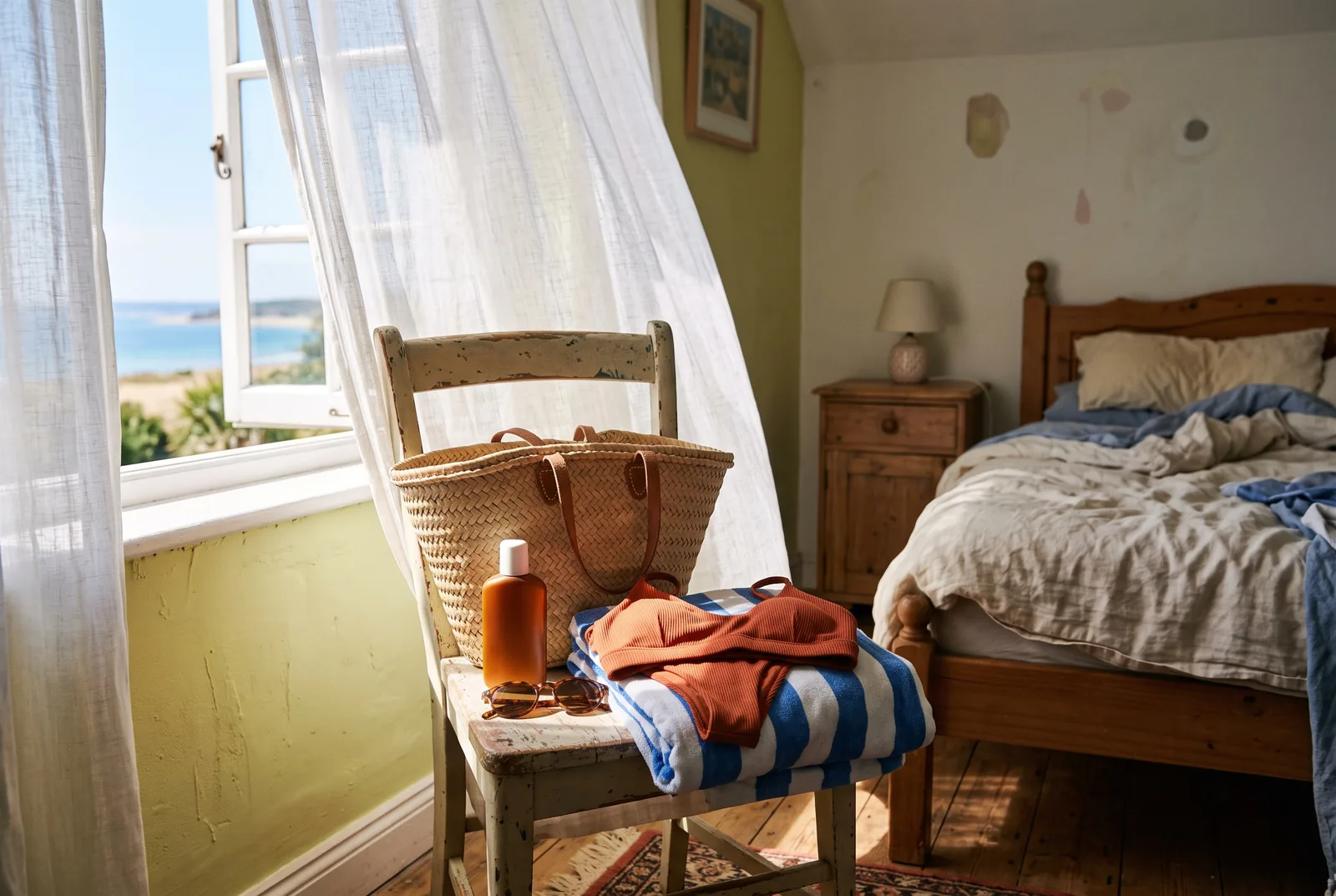 A straw beach bag, striped towel, sunscreen and a folded terracotta bikini on a chair by an open window with the sea outside