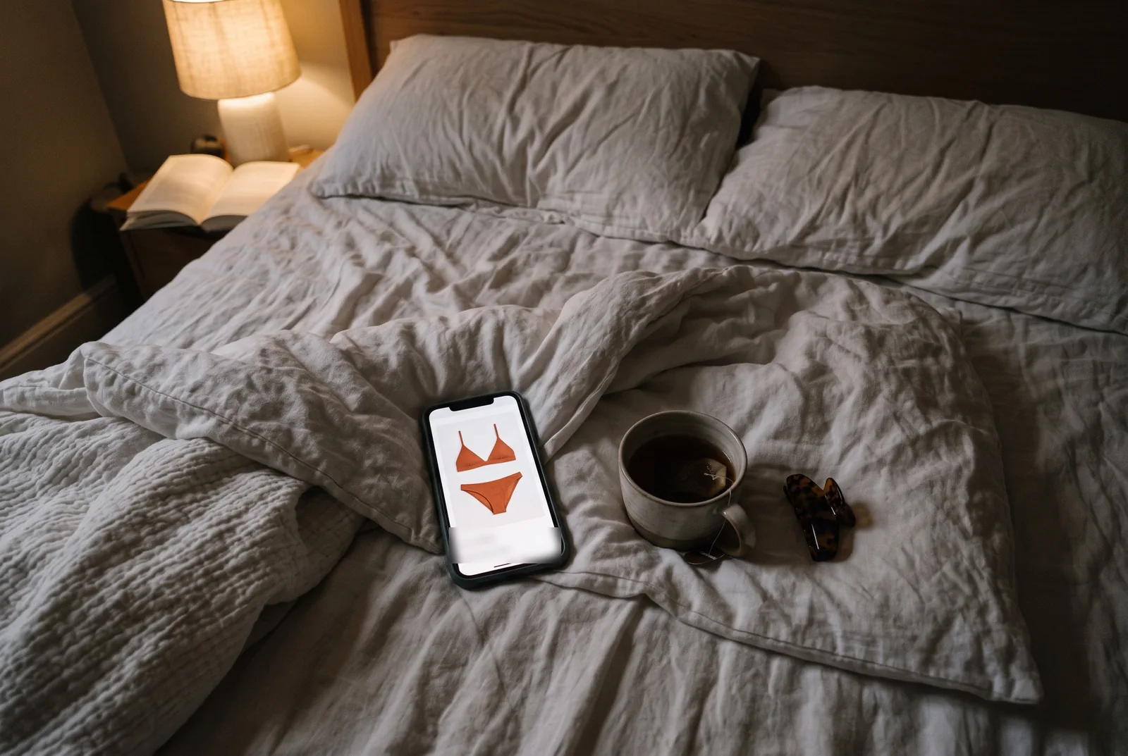 A phone on an unmade bed at night showing a terracotta bikini set on a shop page, next to a mug of tea