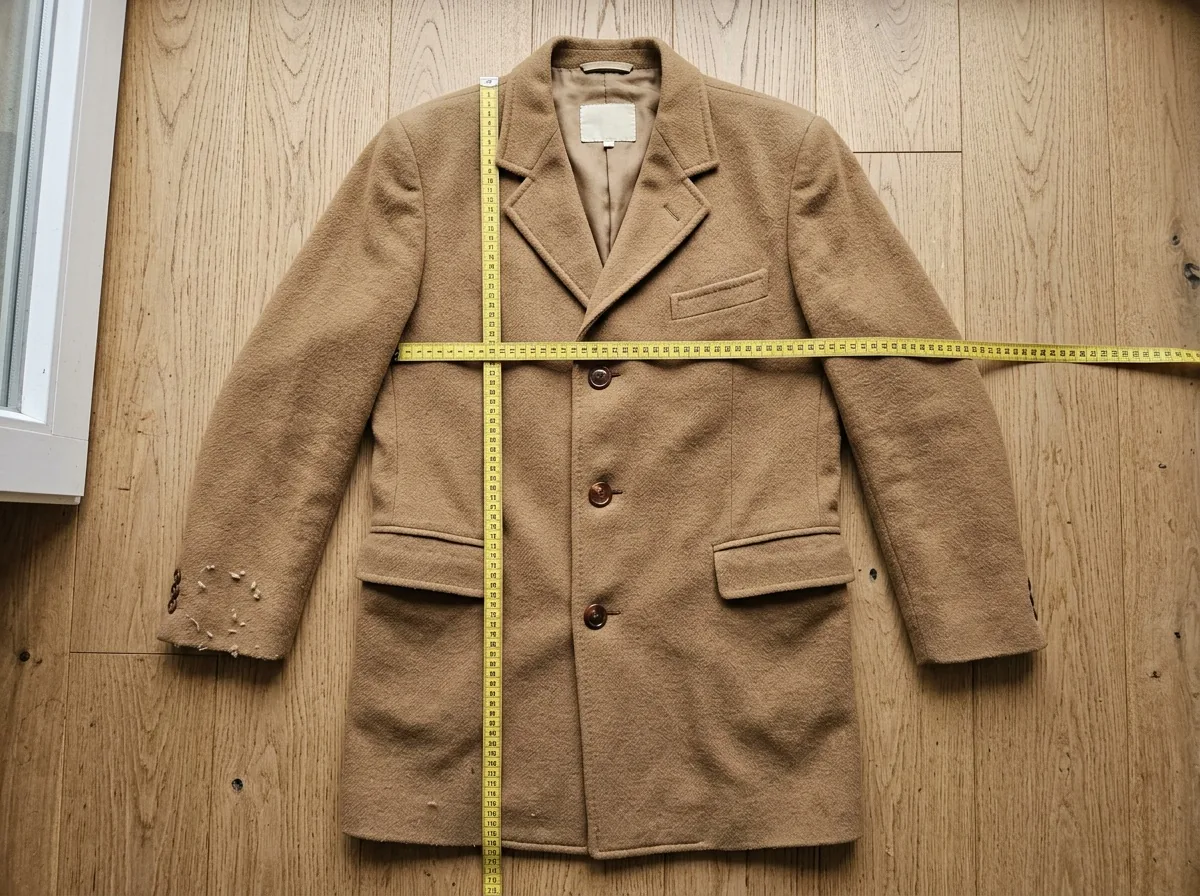 A camel wool coat laid flat on light oak floorboards, buttoned, with a yellow tape measure running from collar to hem and another across the chest, a small cream label at the inside neck and light pilling at one cuff