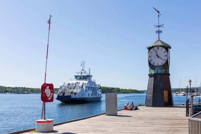 Two people sit in the sun by the clock at Aker Brygge while the ferry passes by