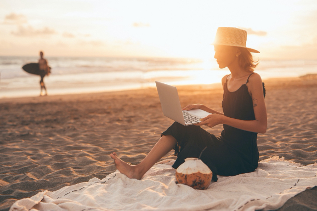 Woman in black dress and hat on blanket at the beach with laptop