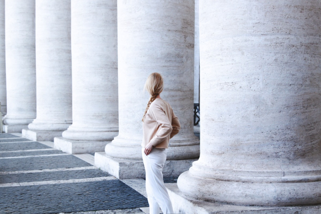 Solitary blonde woman with braid facing away from camera next to a long row of columns