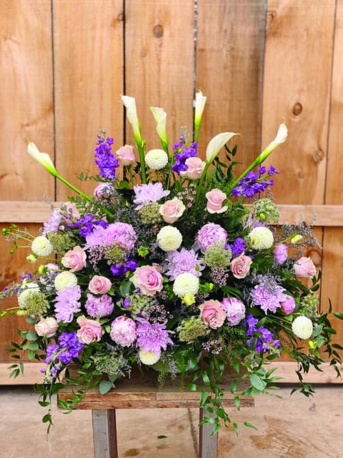 The Watering Can | A large pink and purple side spray made with white calla lilies, soft pink roses, lavender mums, purple stock, soft pink wax flower, purple limonium, white dahlias, chocolate queen anne’s lace, and soft pink peonies backed by greens.
