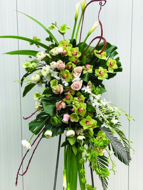 The Watering Can | A stunning modern easel spray made with white calla lilies, bells of Ireland, white & green rununculus, white helleborus, green cymbidium orchids, white dendrobium ochids, light pink roses, fantail willow, and tropical greens.