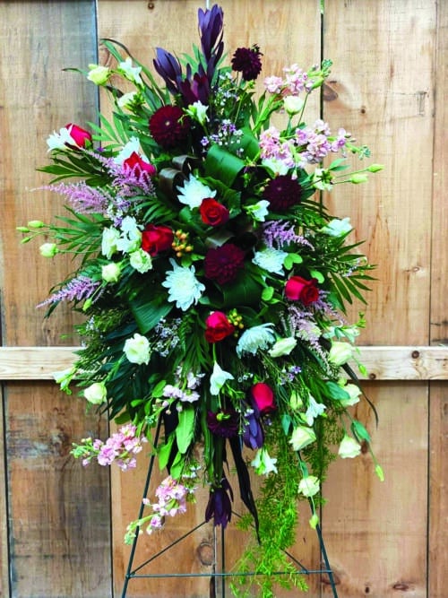The Watering Can | An easel spray made with burgundy leucadendron, soft pink astilbe, burgundy dahlias, red roses, white mums, peach stock, peach hypericum, cream lisianthus, soft pink waxflower, white freesia on a bed of greens.