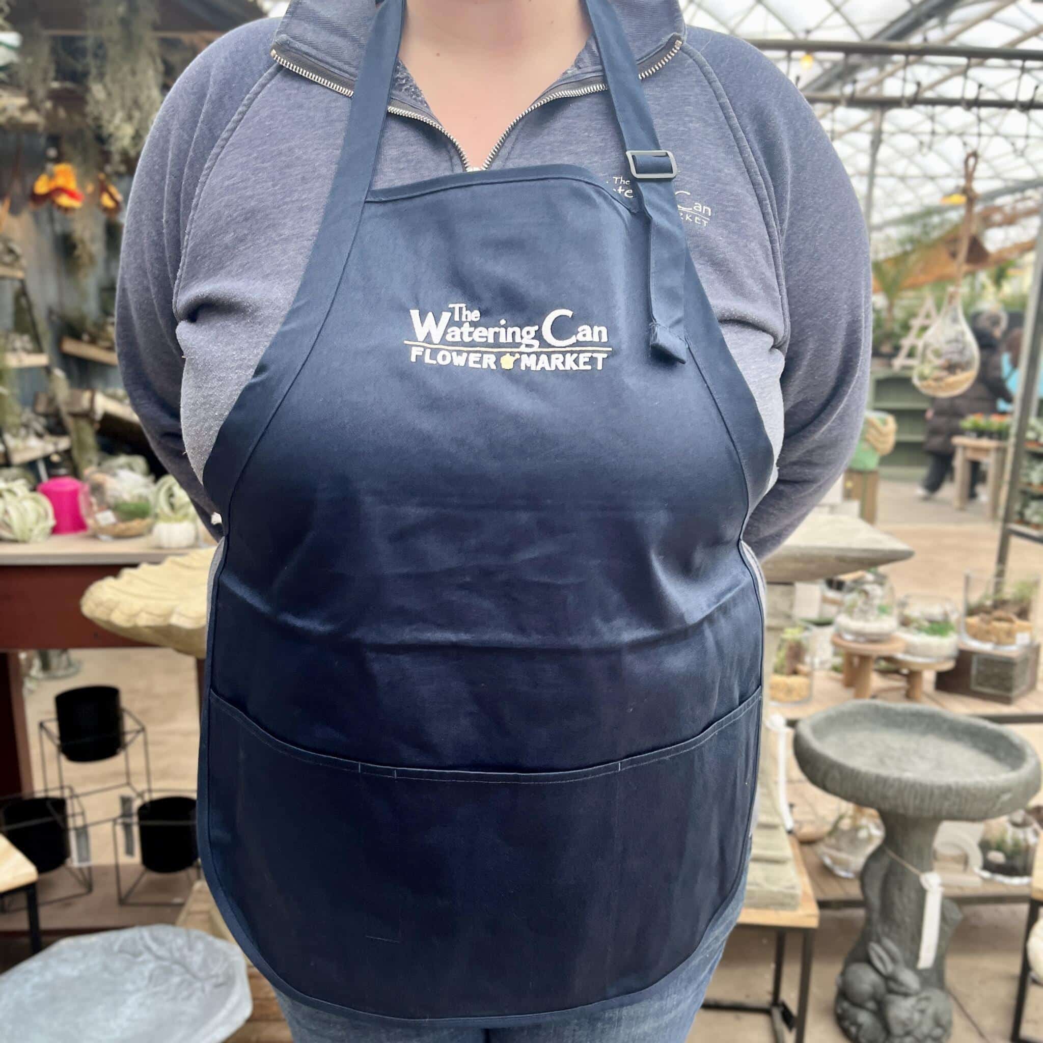 A team member at The Watering Can Flower Market wearing branded gear surrounded by plants and flowers