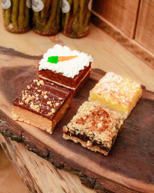 A selection of four assorted baked squares from The Watering Can Pastry Market, showcasing a variety of textures and toppings on a wooden cutting board.