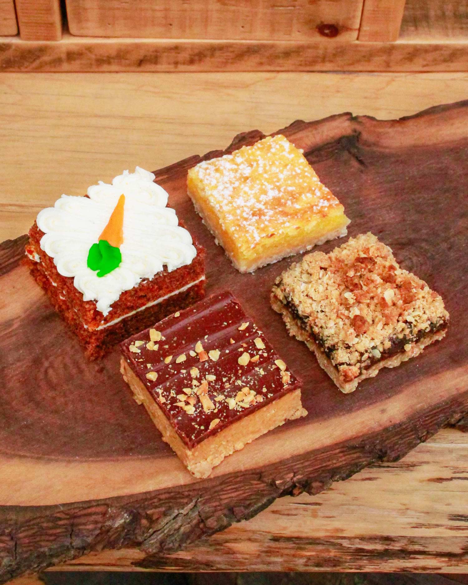 Four assorted pastry squares from The Watering Can Pastry Market, displayed on a wooden board — including rich chocolate and caramel-topped varieties.