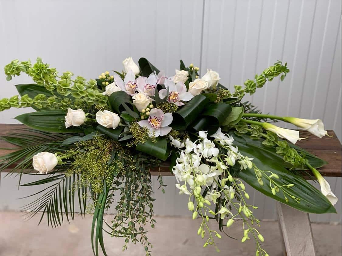 An all white casket spray made with calla lilies, dendrobium orchids, white roses, bells of Ireland, cymbidium orchid blooms, hypericum in a bed of lush greens.