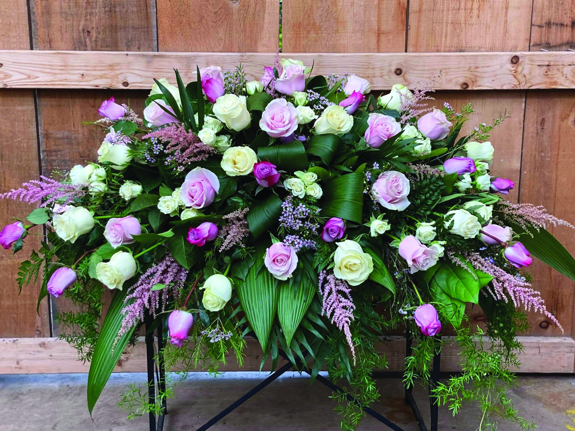 A casket spray made with pink. white, and purple roses alongside astibille, waxflower and lush greens.