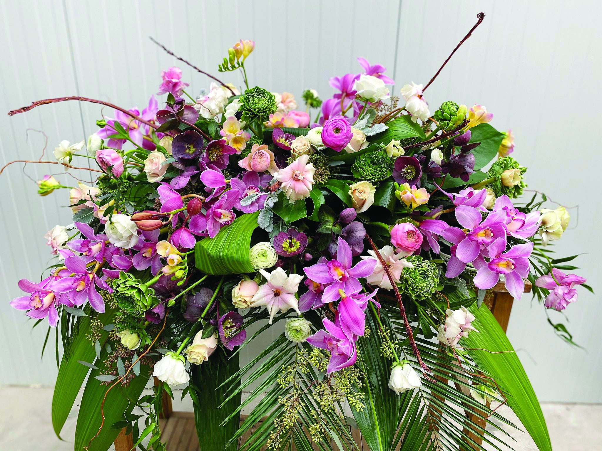 A large and luxurious purple casket spray made with pink cymbidium orchid, pink roses, soft pink freesia, dark purple helleborus, ranunculus, butterfly ranunculus, and white anemone on a lush bed of tropical greenery.