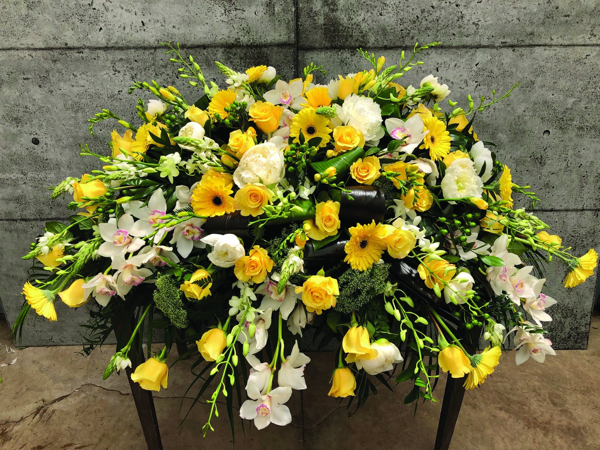 A yellow casket spray made with white cymbidium orchid, bright yellow roses, white peonies, bright yellow gerberas, white dendrobium orchids, stars of Bethlehem, yellow freesia, green hypericum, and white trachelium on a bed of greens.