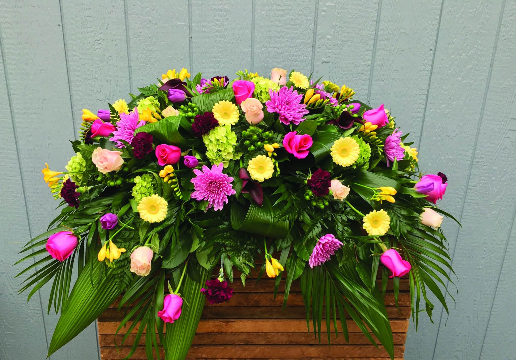 A pink and yellow casket spray made with hot pink roses, soft pink roses, lavender mums, lime green hydrangea, dark purple moon carnations, dark pink calla lilies, pink tulips, yellow freesia, green hypericum, and soft yellow gerbera on a lush bed of greens.
