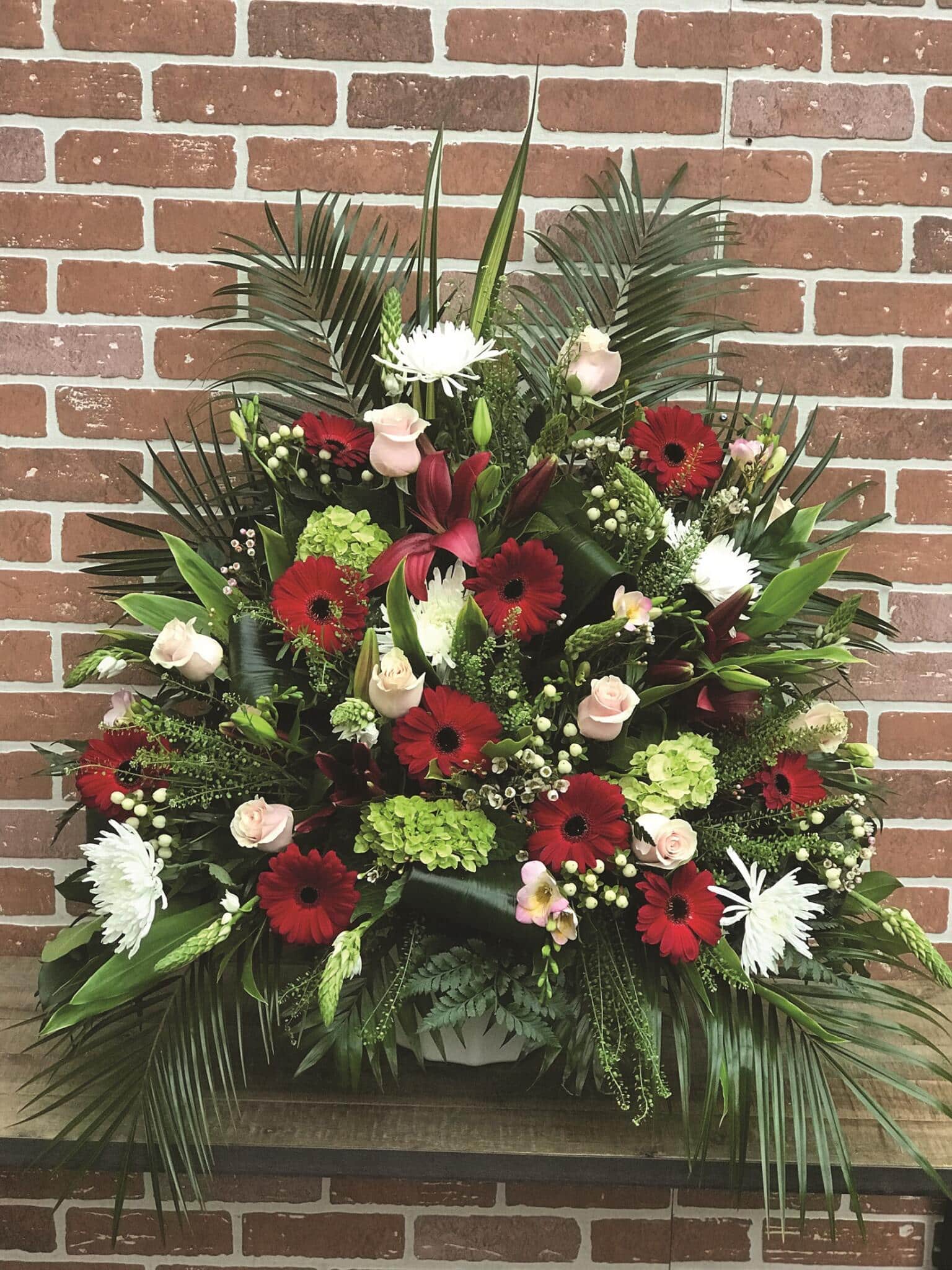 A large side spray made with soft pink roses, white mums, burgundy lilies, burgundy gerberas, green hydrangea, stars of Bethlehem, white hypericum, soft pink freesia, white waxflower backed by greenery.