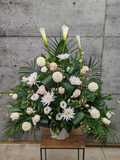 An all white side spray made with calla lilies, dahlias, roses, waxflower, lisianthus, and mums backed by greens.