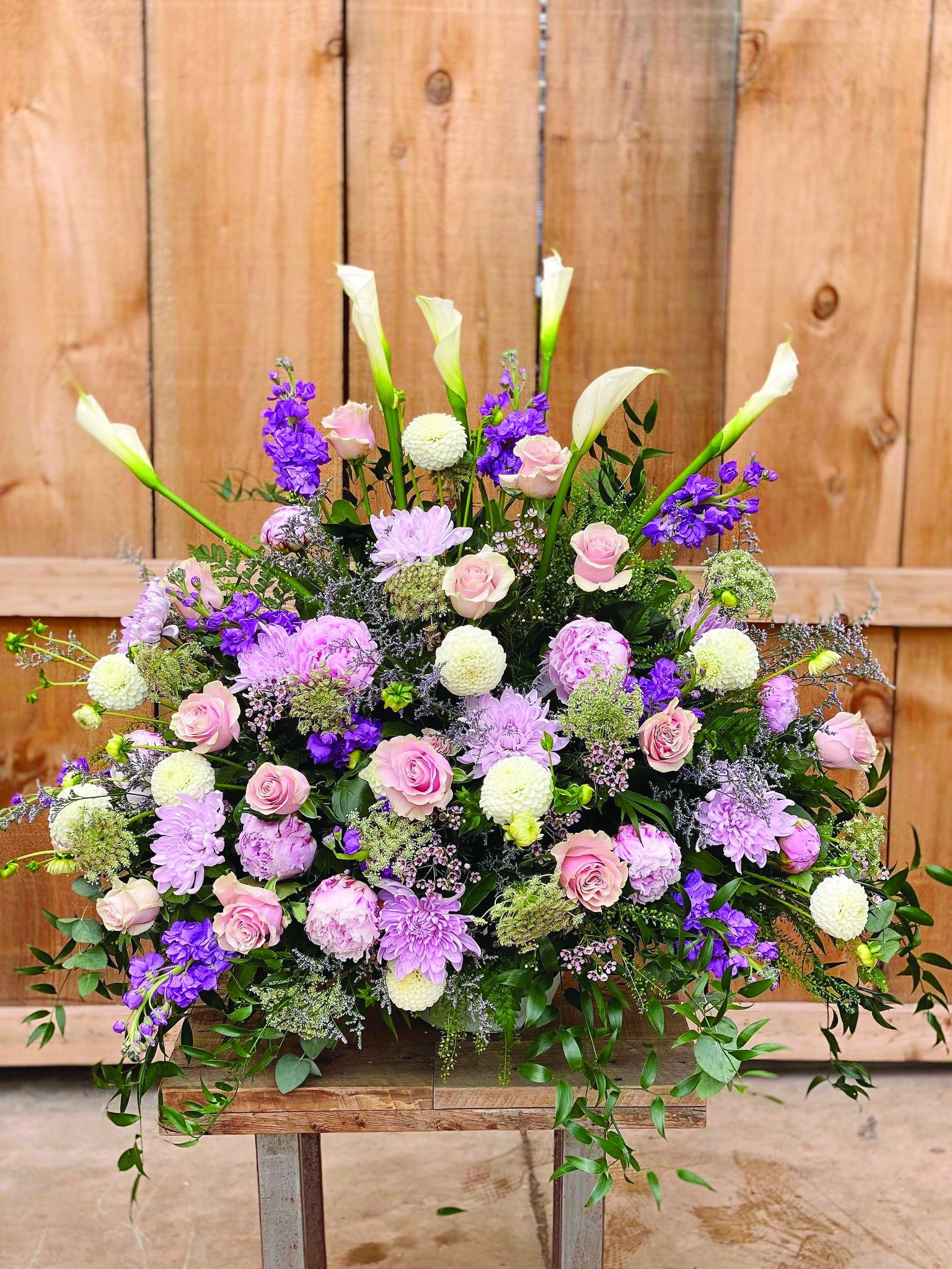 A large pink and purple side spray made with white calla lilies, soft pink roses, lavender mums, purple stock, soft pink wax flower, purple limonium, white dahlias, chocolate queen anne’s lace, and soft pink peonies backed by greens.