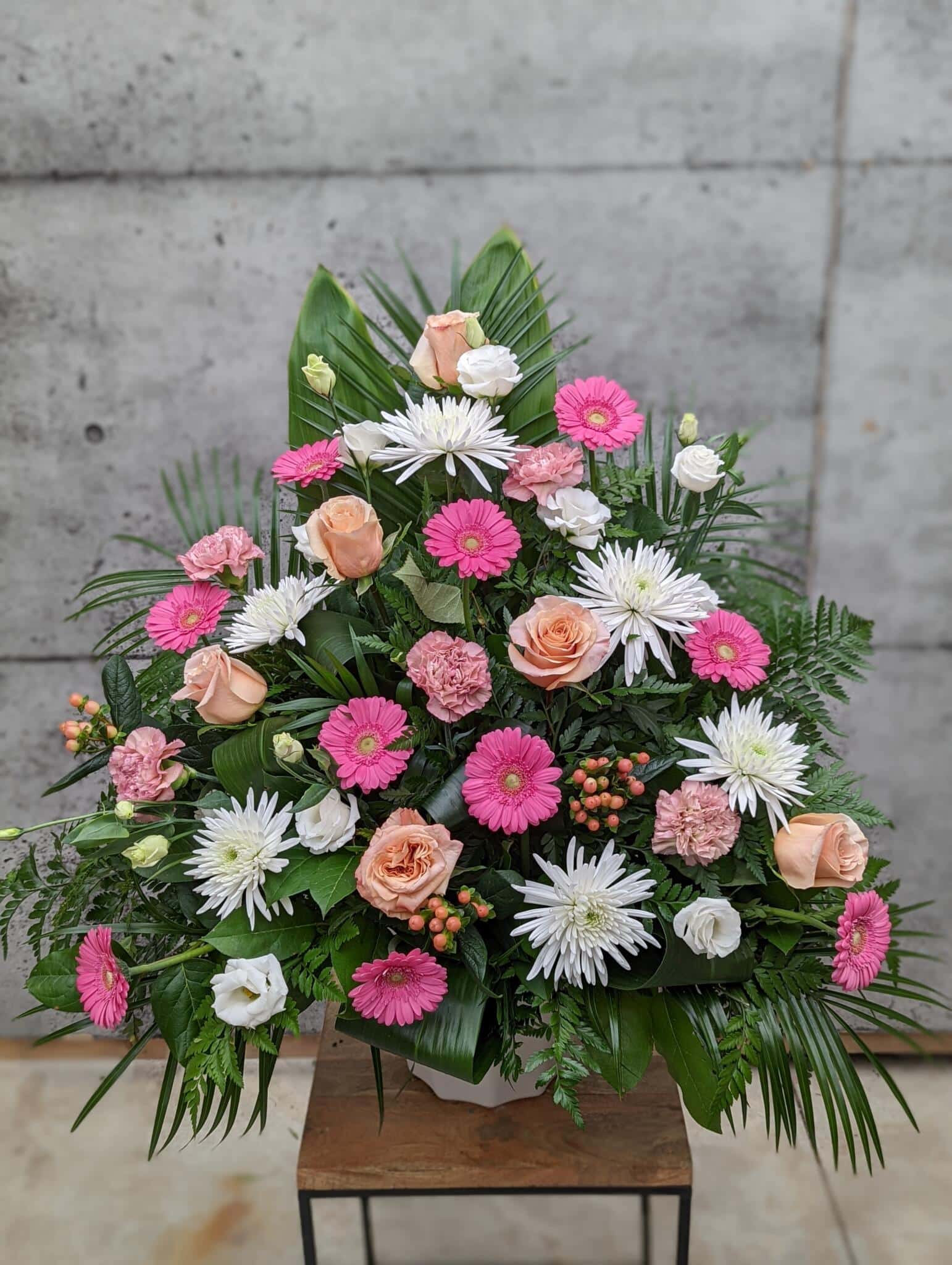 A large peach, pink, and white side spray made with white mums, shimmer roses, pink gerberas, peach hypericum, dusty pink carnations, and white lisianthus backed by greens.