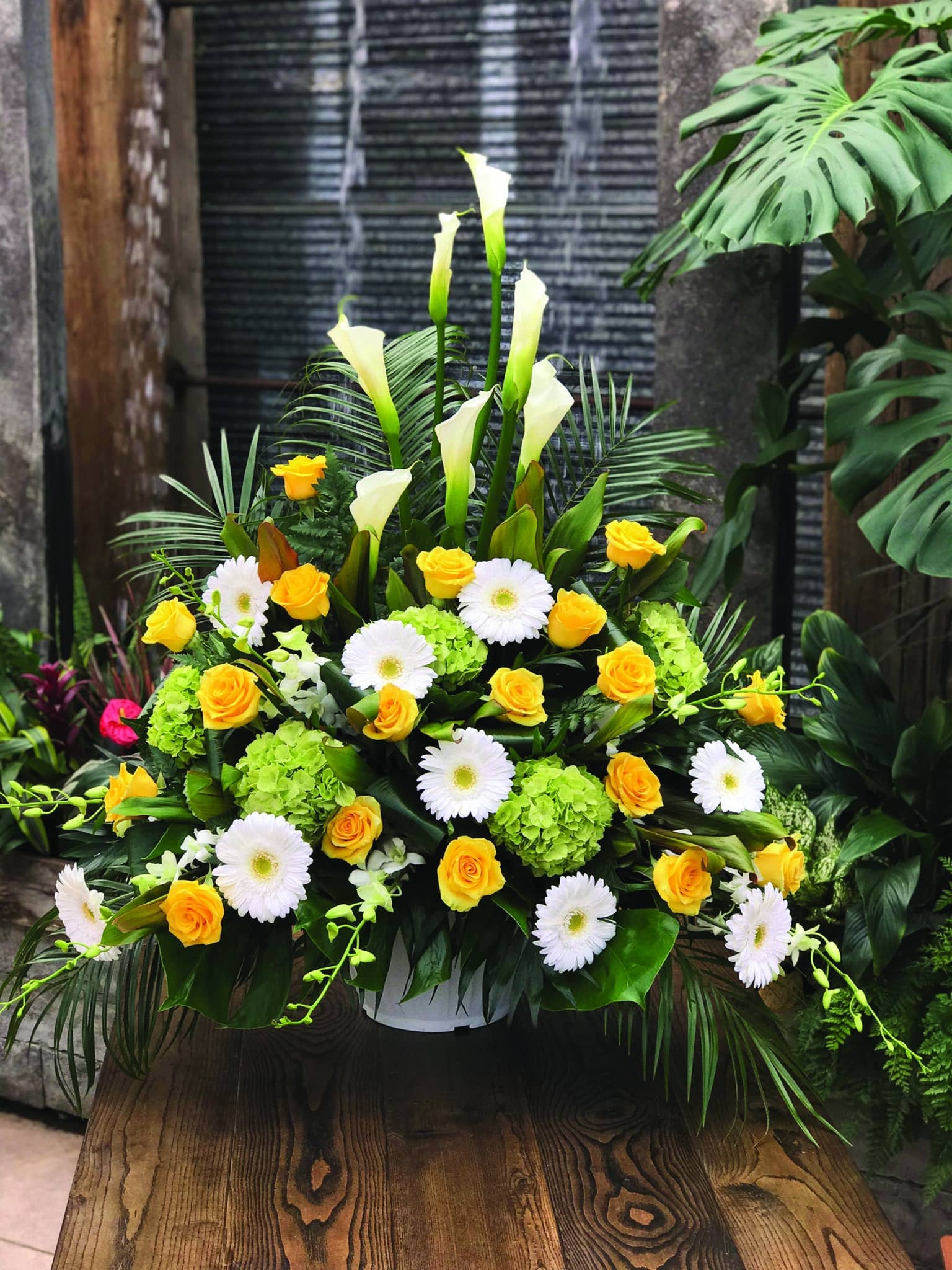 A yellow, white, and green side spray made with white calla lilies, green hydrangea, yellow roses, white gerberas, and white dendrobium orchids backed by greenery.
