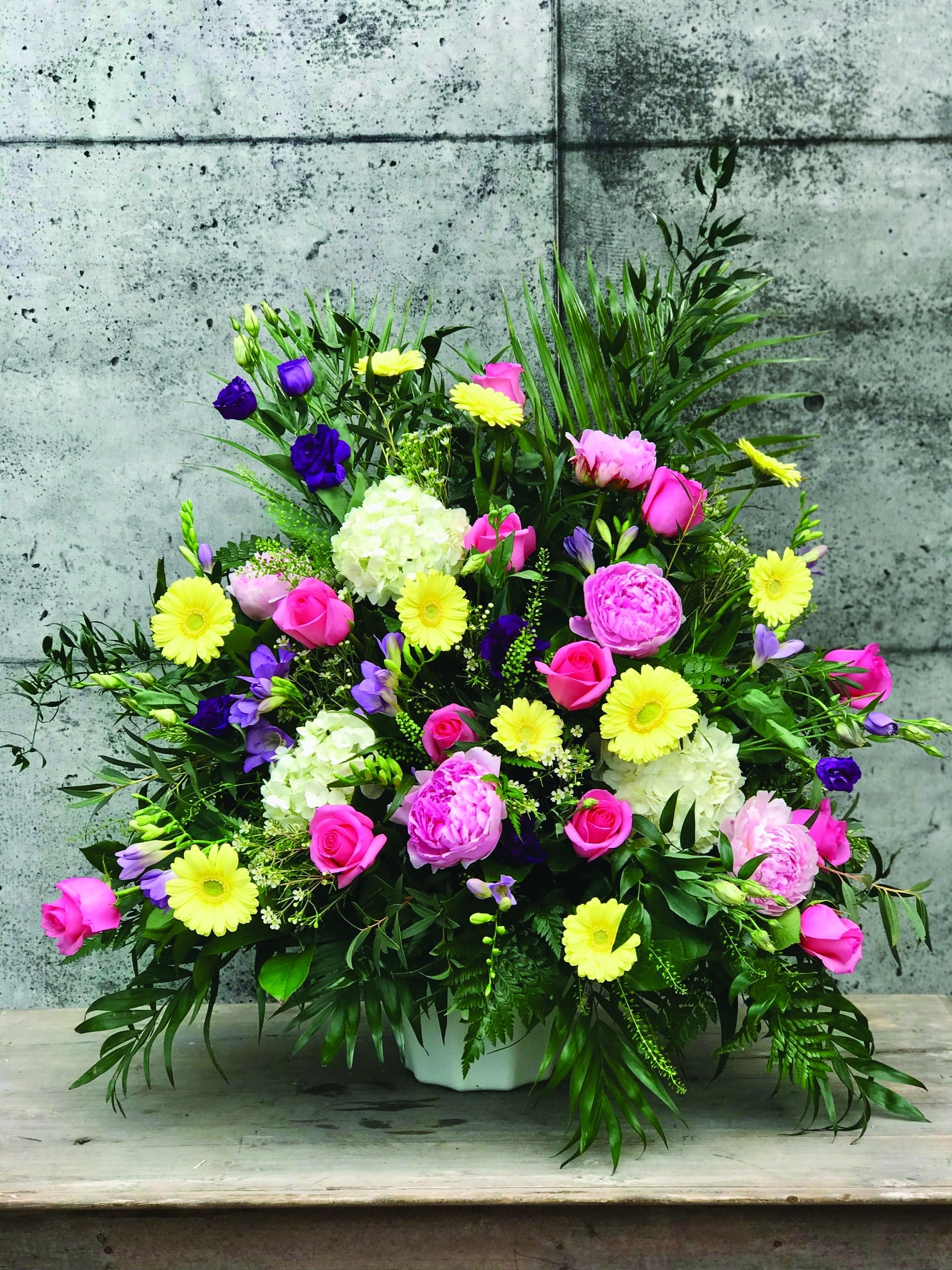 A pink and yellow side spray made with white hydrangea, pink roses, lavender freesia, purple lisianthus, white waxflower, yellow gerbera daisies, and soft pink peonies backed by a variety of greens.