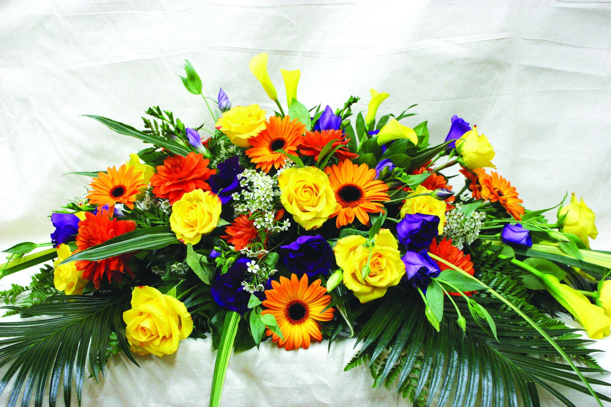 A colourful casket spray made with yellow calla lilies, yellow roses, orange gerberas, orange dahlia, purple lisianthus, white wax flower, and purple purple freesia on a bed of greens.