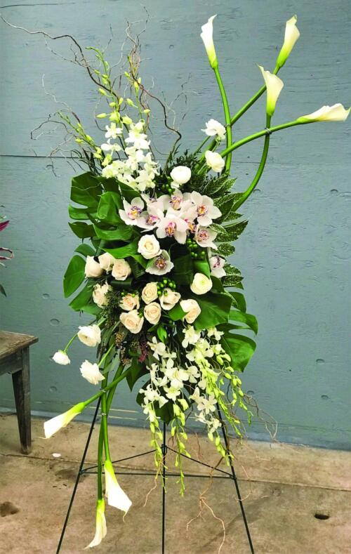 An elegant white easel spray made with white calla lilies, white roses, stem white cymbidium orchid, white dendrobium orchids, green hypericum, white ranunculus, and tropical greens.