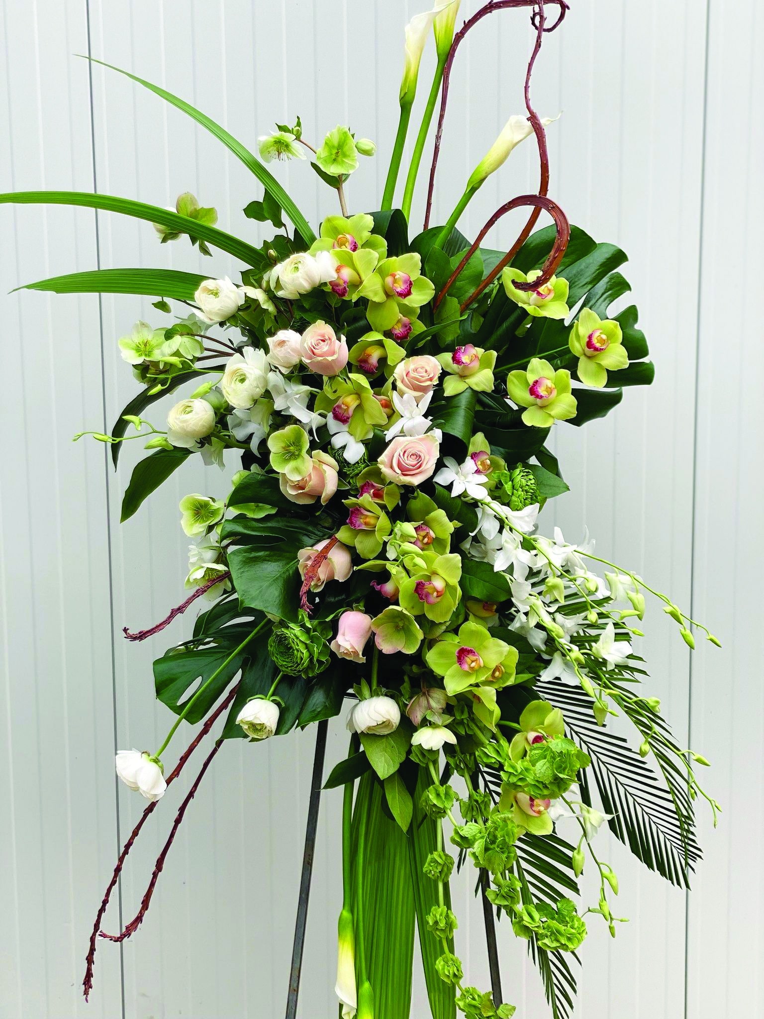 A stunning modern easel spray made with white calla lilies, bells of Ireland, white & green rununculus, white helleborus, green cymbidium orchids, white dendrobium ochids, light pink roses, fantail willow, and tropical greens.