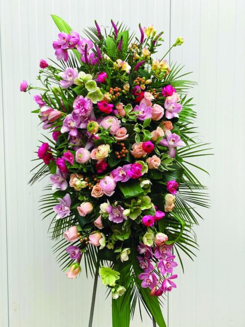 A large pink easel spray made with roses, helleborus, butterfly rununculus, rununculus, hypericum, cymbidium orchid, freesia, and veronica on a bed of greens.