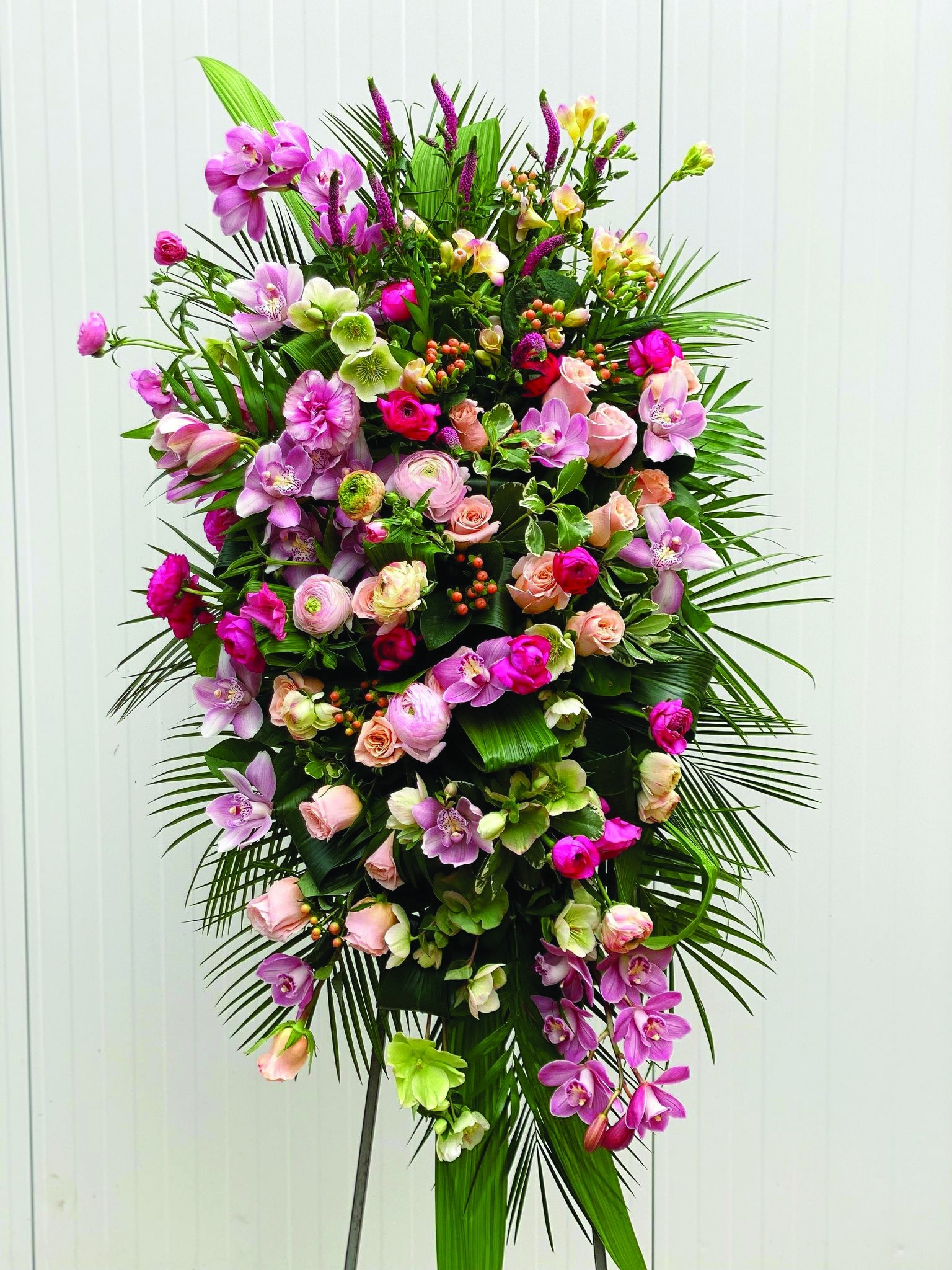 A large pink easel spray made with roses, helleborus, butterfly rununculus, rununculus, hypericum, cymbidium orchid, freesia, and veronica on a bed of greens.