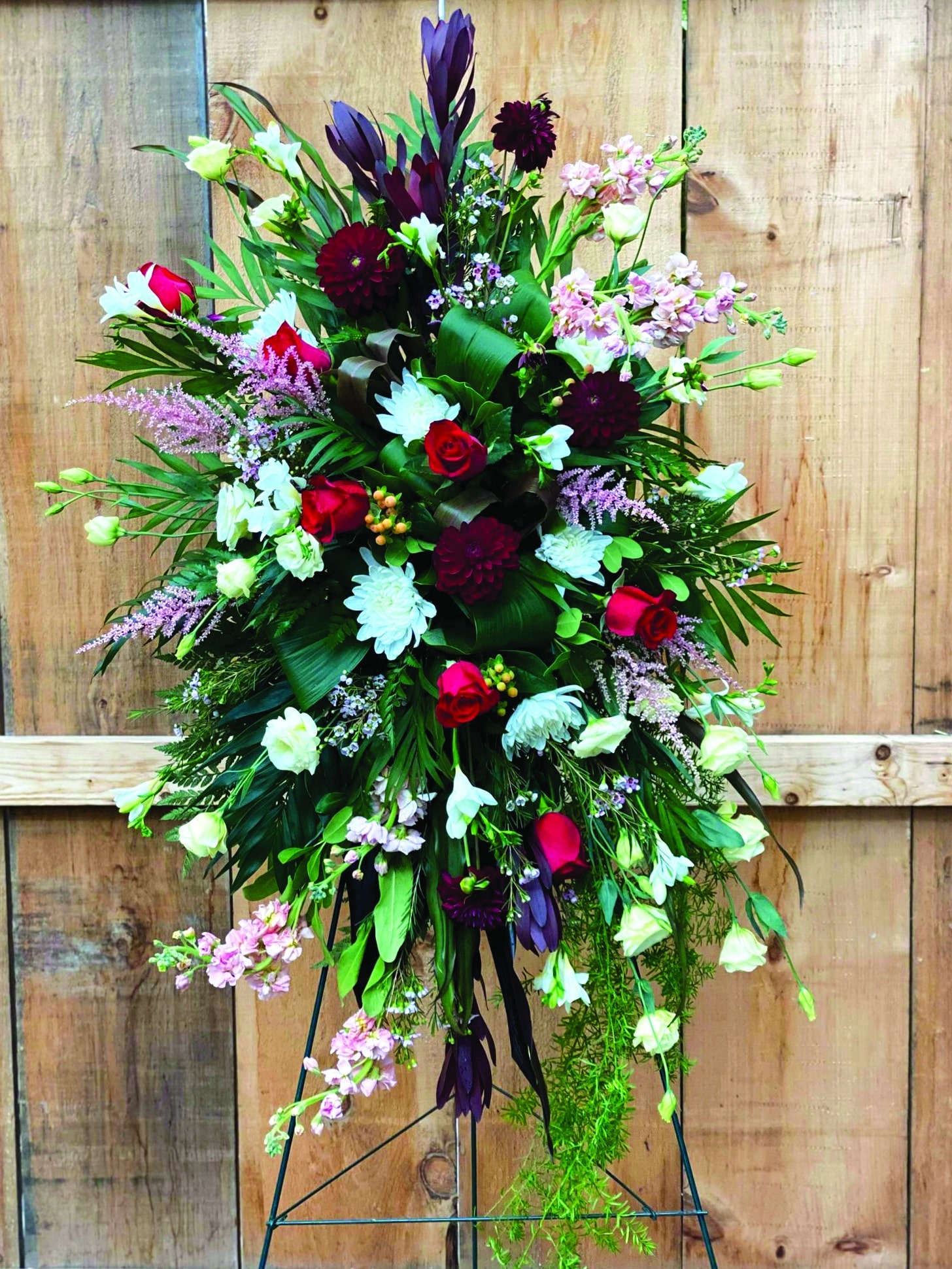 An easel spray made with burgundy leucadendron, soft pink astilbe, burgundy dahlias, red roses, white mums, peach stock, peach hypericum, cream lisianthus, soft pink waxflower, white freesia on a bed of greens.