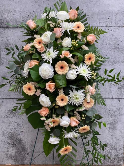 A peach and white easel spray made with roses, mums, rununculus, hypericum, statice, and gerberas on a lush bed of greens.